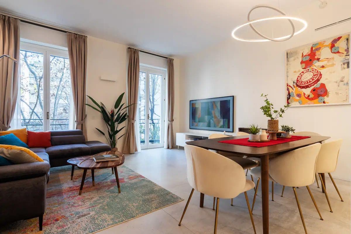 Open space con divano grigio, tavolo da pranzo, lampadario ad anello e finestre su strada alberata di Brera