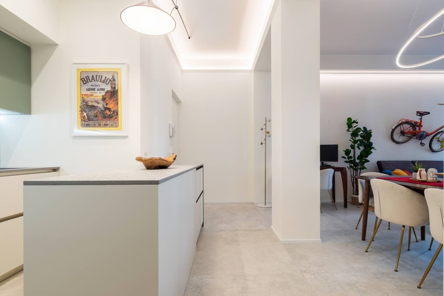 Vista dalla cucina verso il living con isola bianca, poster vintage Braulio e illuminazione LED a soffitto