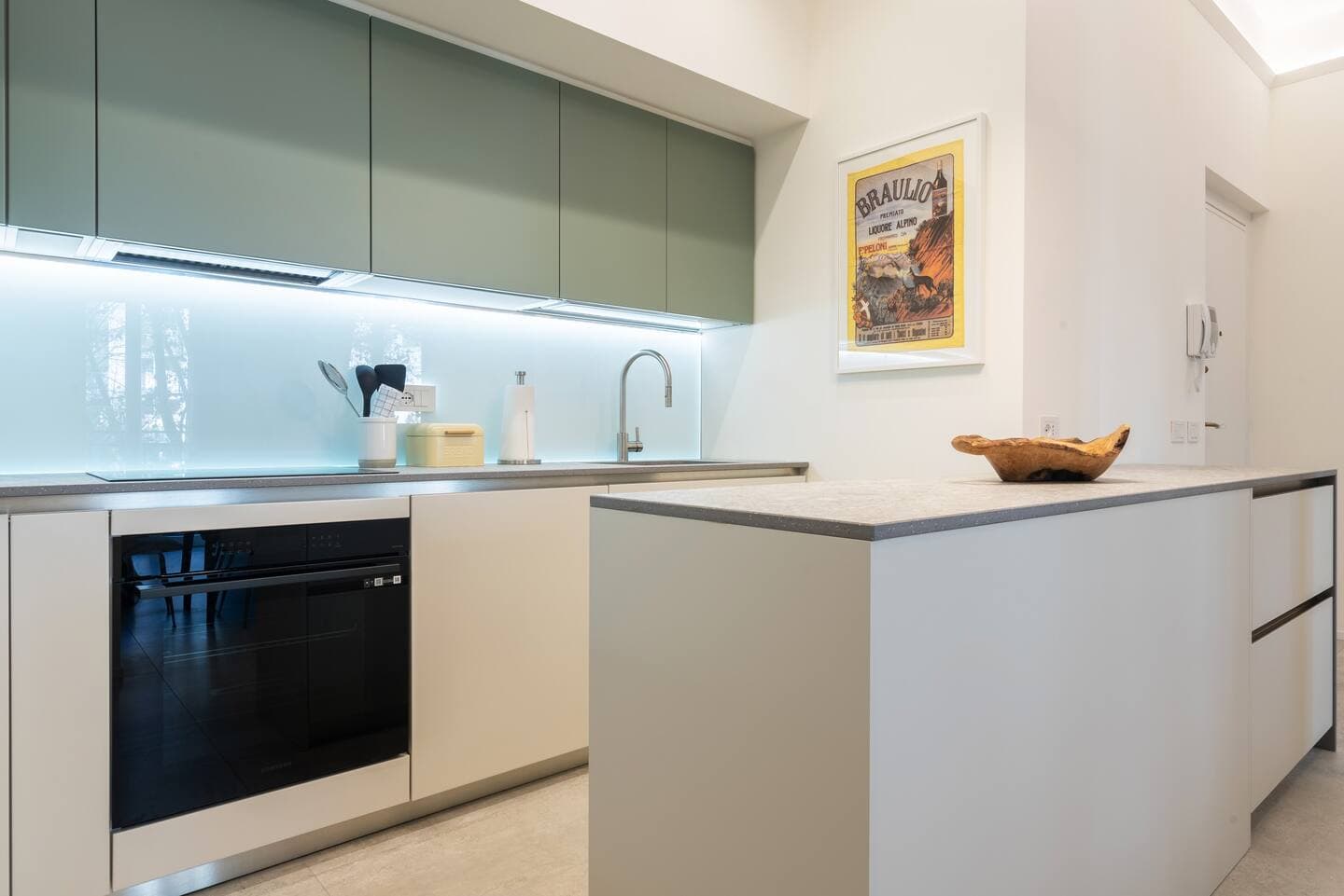 Cucina con pensili verde salvia retroilluminati, forno incassato nero, piano in acciaio e poster Braulio