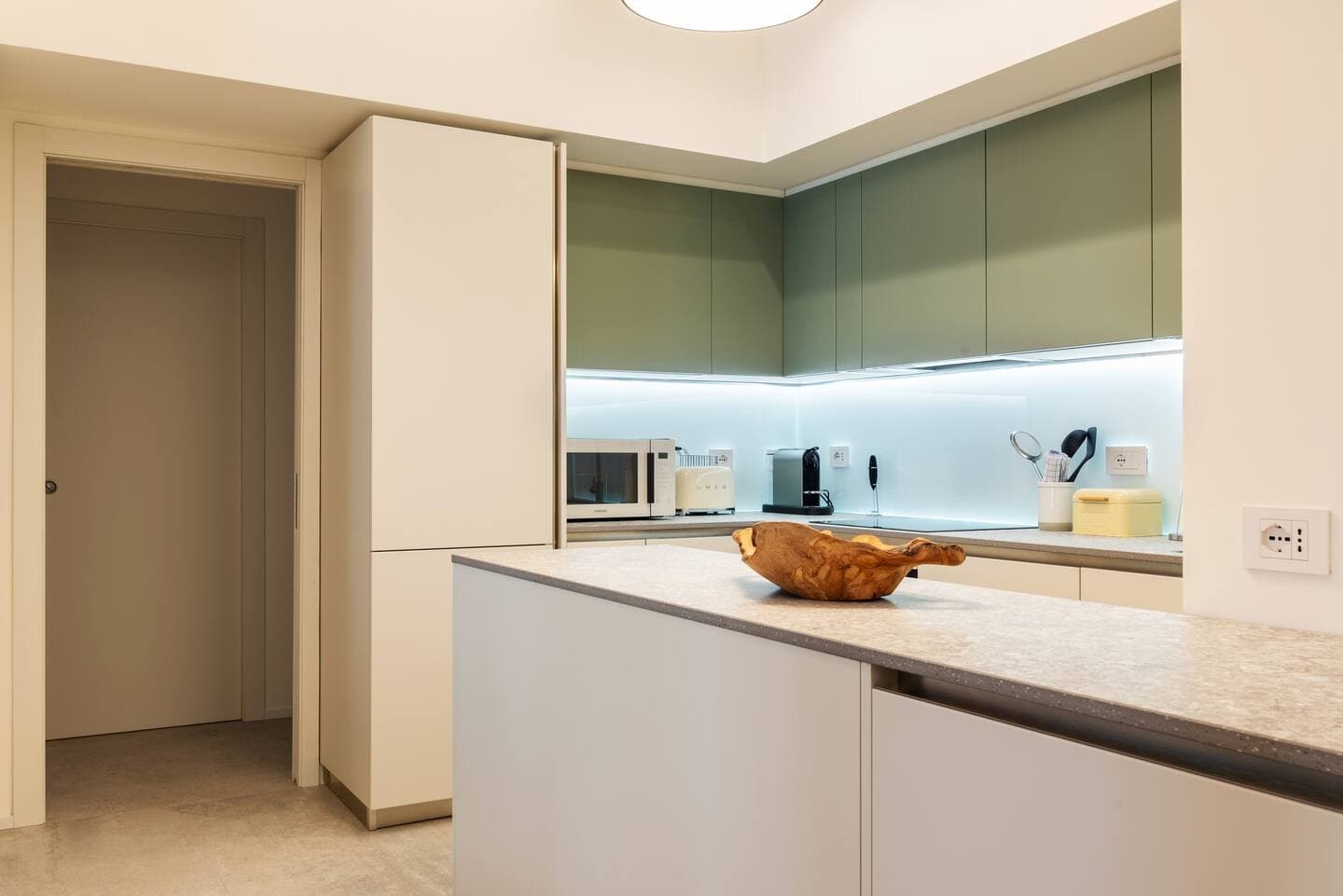 Cucina con isola, pensili verde salvia retroilluminati, piccoli elettrodomestici e vista sul corridoio