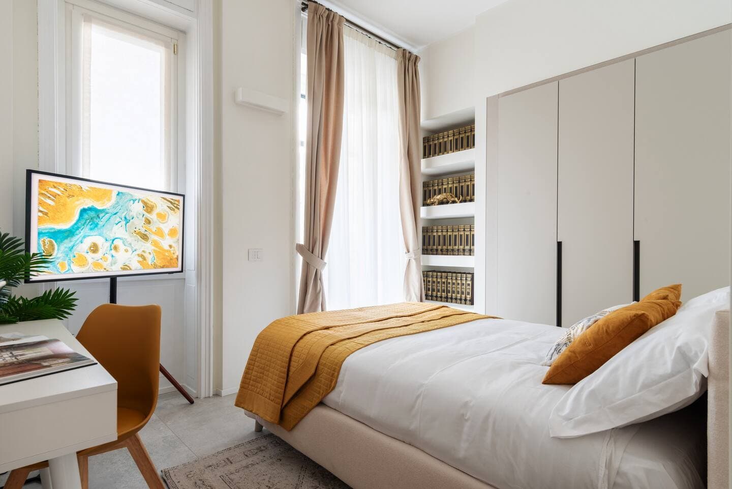 Terza camera con letto e plaid senape, scrivania con Samsung Frame TV su cavalletto e libreria con volumi dorati