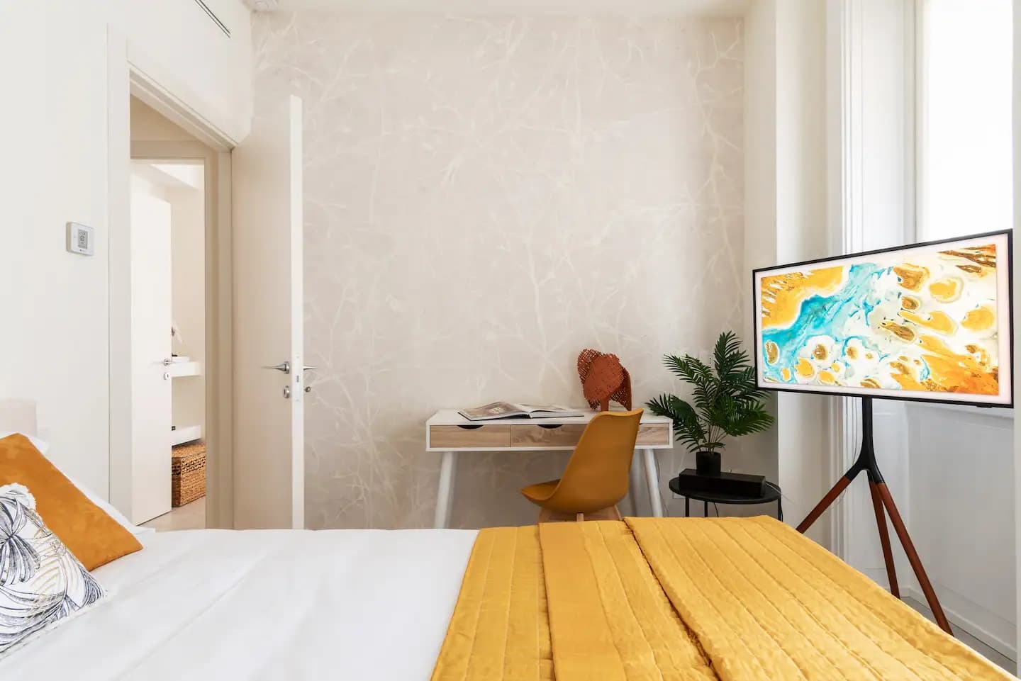 Terza camera con scrivania, sedia senape, Samsung Frame TV su cavalletto e parete texturizzata beige