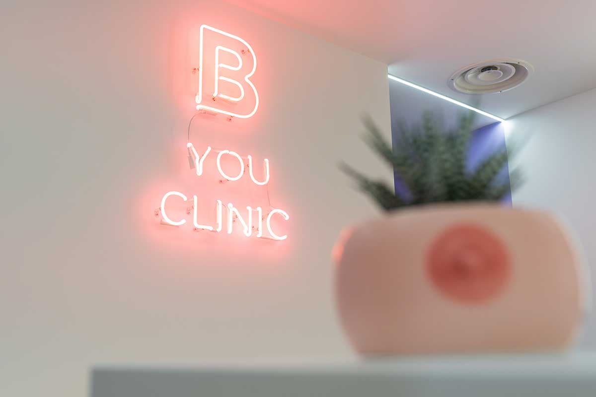 Insegna neon rosa B-You Clinic su parete bianca con vaso decorativo in primo piano