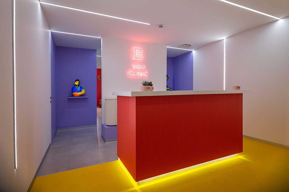 Reception con bancone rosso, illuminazione LED gialla, neon B-You Clinic e pareti colorate