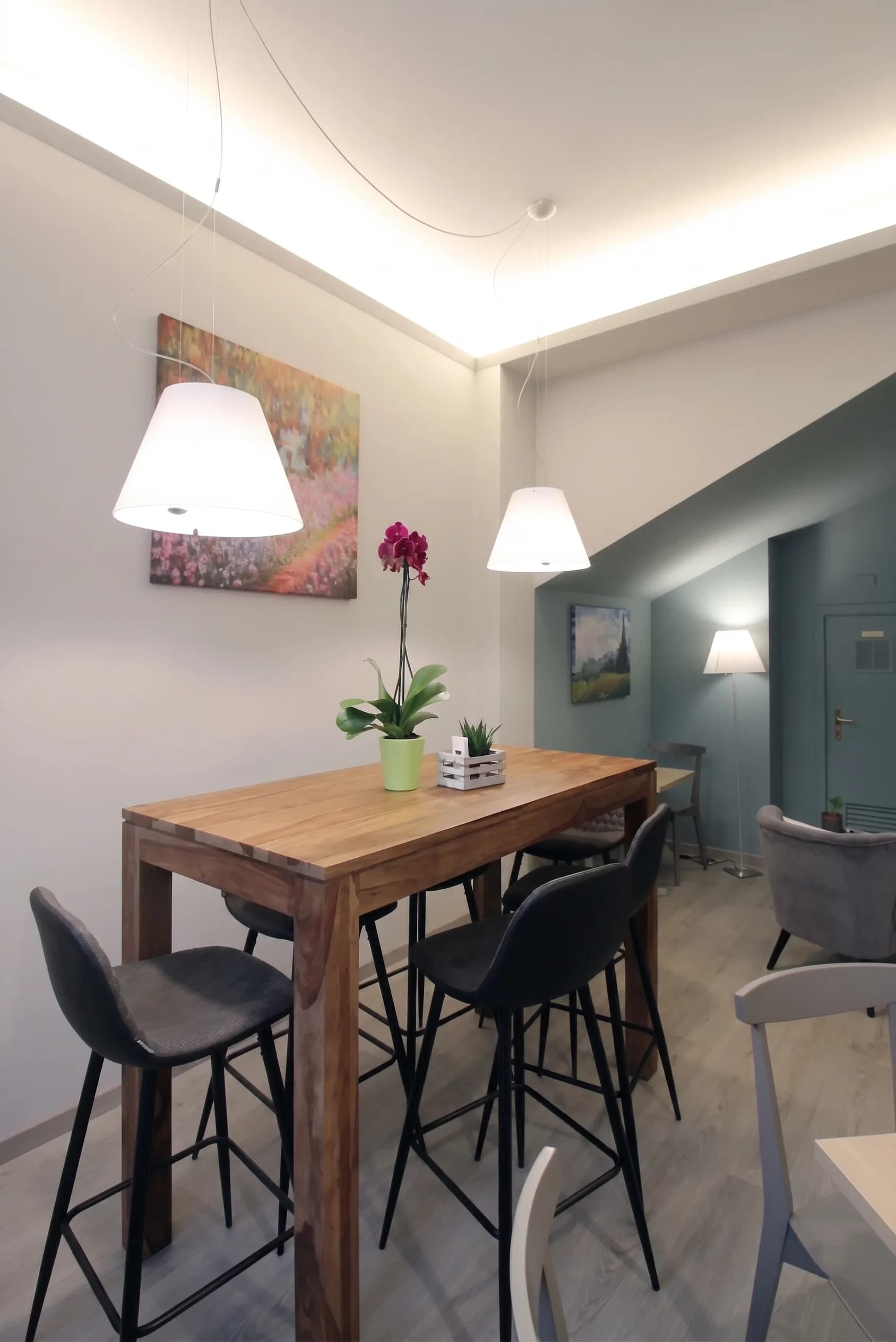 Tavolo alto in legno con sgabelli, lampade a sospensione bianche e quadro impressionista