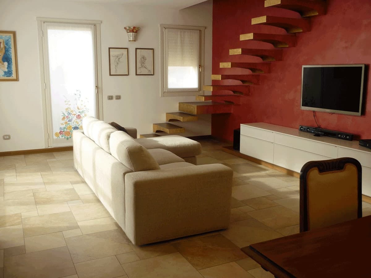 Soggiorno con divano beige, scala a sbalzo su parete rossa, TV e quadri a parete